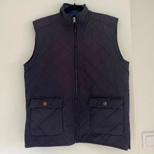 Quiltad mörkblå väst View Horse Unique - Quiltad mörkblå väst från View Horse Unique med dragkedja framtill och två stora fickor med lock och knapp. Insidan är fodrad med blå fleece för extra värme. 