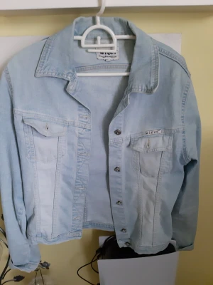 Ljusblå jeansjacka från Wiges - Säljer en klassisk ljusblå jeansjacka från Wiges i storlek S. Jackan har två bröstfickor med knappar, silverfärgade knappar framtill och en avslappnad passform. Perfekt att slänga över en hoodie eller t-shirt för en chill look.