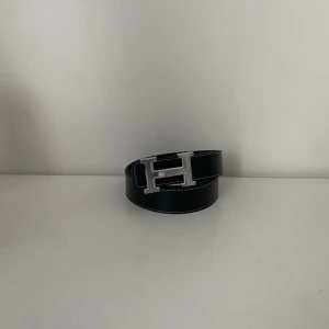 HermesBälte - Snyggt svart skärp med ett stort H-format spänne i silverfärg. Skärpet har vita sömmar längs kanterna som ger en schysst kontrast. Perfekt accessoar för att lyfta din outfit med en stilren detalj.Längd 110cm