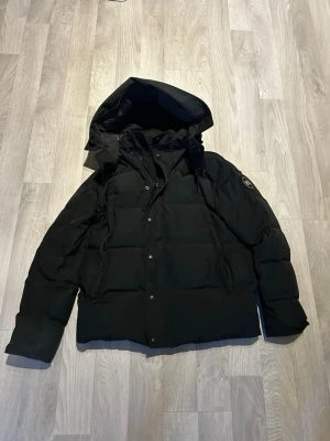 Svart dunjacka från Canada Goose - Svart dunjacka från Canada Goose med huva och snyggt märke på ärmen. Jackan har knappar och dragkedja framtill, samt två sidofickor. Materialet är polyester och bomull på utsidan, med dunfyllning för extra värme. Perfekt för kalla dagar och har en klassisk, stilren look. Päls och påse ingår 
