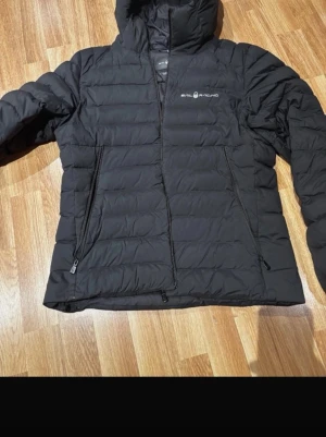 Svart pufferjacka från Sail Racing - Säljer en svart pufferjacka från Sail Racing med huva och dragkedja framtill. Jackan har två sidofickor med dragkedja och är quiltad för extra värme. Perfekt för kalla dagar och har en stilren look som funkar till det mesta.