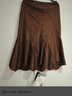 Brun långkjol med volangkant - Säljer en brun långkjol i storlek L med snygg volangkant nedtill. Kjolen har en glansig yta och ett elegant fall som ger en cool och trendig look. Perfekt att styla med boots eller sneakers för en chill vibe.