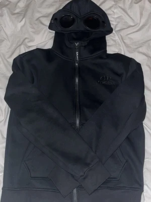 C.P. Company hoodie med goggles – svart  - Svart hoodie från C.P. Company med ikoniska goggles i huvan. Klassisk zip-modell med fickor framtill och logotyp på bröstet. Mjuk och varm, perfekt för vardagsbruk. Använd men i fint skick, inga större defekter. Äkta vara – se etikett på bild. Passar både streetwear och casual stil.