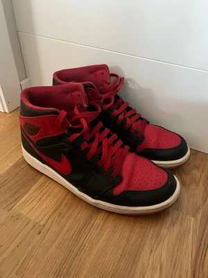 Nike Air Jordan 1 High röd/svart - Säljer ett par Nike Air Jordan 1 High sneakers i klassisk röd och svart färg med vita sulor. Skorna har snörning, perforerad tå och ikonisk swoosh på sidan. Tillverkade i skinn med högt skaft och rund tå. Perfekta för dig som gillar streetstyle och vill sticka ut.