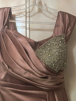 Champagnefärgad långklänning med paljetter - Säljer en elegant långklänning i champagnefärgad satin med breda axelband och glittrig paljettdekor över ena bröstet. Klänningen har draperad överdel och en snygg slits framtill som ger extra rörelse. Perfekt för bal eller fest.