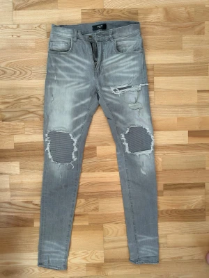 Grå skinny jeans från Amiri - Säljer ett par grå skinny jeans från Amiri med slitna detaljer och stora hål vid knäna med ribbad tyg under. Jeansen har fem fickor, dragkedja och knapp i midjan. Färgen är ljusgrå med tvättad look och de har låg midja.
