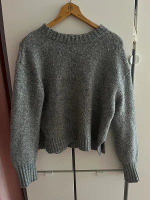 Grå stickad tröja med ribbade muddar - Mysig grå stickad tröja med ribbad rund hals och muddar vid ärmslut och nederkant. Tröjan har en loose passform och är perfekt för kyliga dagar. Enkel och stilren design som passar till jeans eller mjukisbyxor.