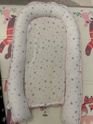 Rosa och vit babyväst med stjärnor - Säljer en mysig babyväst i ljusrosa och vit med gråa stjärnor. Västen har en mjuk, bubblig yta i rosa syntetmaterial och en avtagbar vit del med stjärnmönster. Perfekt för att ge extra komfort och stil till de minsta.