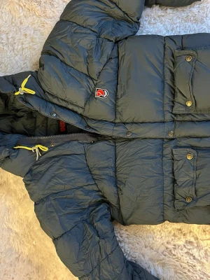 Fjällräven jacka - Säljer en grå/mörkblå pufferjacka från Fjällräven med gul snörning och huva. Jackan har stora fickor med knappar, dragkedja och Fjällräven-logga på bröstet. Perfekt för kalla dagar!