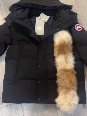 Svart dunjacka från Canada Goose - Svart dunjacka från Canada Goose med huva och avtagbar pälsdetalj i beige och bruna toner. Jackan har klassisk patch på ärmen, knappar och dragkedja framtill. Perfekt för kalla dagar och har en schysst, tidlös look.