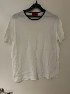 Vit t-shirt från Hugo Boss - Säljer en stilren vit t-shirt från Hugo Boss med rund hals och diskret svart kant runt kragen. T-shirten har korta ärmar och är gjord i mjuk bomull som känns skön mot huden. Perfekt för dig som gillar enkel och clean stil.