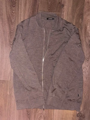 Brun cardigan från J.Lindeberg i merinoull - Säljer en brun cardigan från J.Lindeberg i storlek M, tillverkad i 100% merinoull. Koftan har lång ärm, ribbade muddar och dragkedja framtill. Perfekt för dig som gillar stilrena och bekväma plagg med klassisk look.