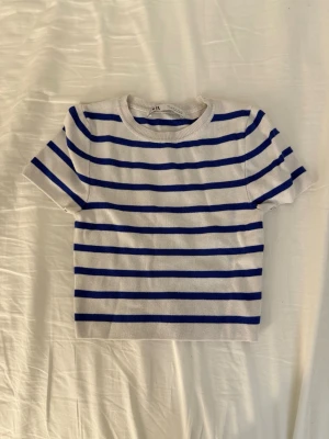 Randig topp från Zara i blått och vitt - Säljer en kortärmad topp från Zara i storlek S. Toppen är vit med horisontella marinblå ränder och har en rund halsringning. Toppen har en tight passform. Materialet är mjukt och stickat, vilket gör den skön att bära. Perfekt för dig som gillar klassisk och enkel stil. 