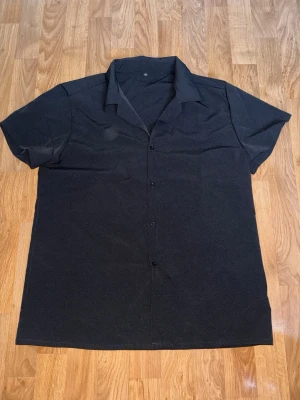 Svart kortärmad skjorta med knappar - Snygg svart skjorta med korta ärmar och klassisk krage. Skjortan har knäppning framtill och en clean, enkel design utan mönster eller extra detaljer. Perfekt för dig som gillar stilrena plagg och vill ha något som funkar till många olika outfits.