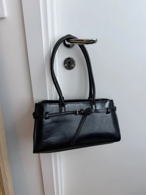 Svart handväska med spänne Stradivarius  - Stilren svart handväska i skinnimitation med dubbla handtag och dekorativt bälte framtill. Väskan har ett rymligt huvudfack, innerficka med dragkedja och avtagbar axelrem. Perfekt till dig som gillar klassisk och clean look. Oanvänd 