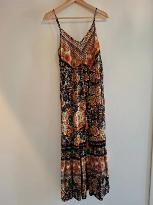 Blommig långklänning Rusttydusty - Säljer en bohemisk långklänning från Rusttydusty i storlek L. Klänningen har tunna axelband, blommigt och mönstrat tryck i orange, svart, vit och gult med inslag av hästar. Materialet är mjukt och lätt, perfekt för varma dagar. Volangdetaljer och dekorativa tofsar framtill.