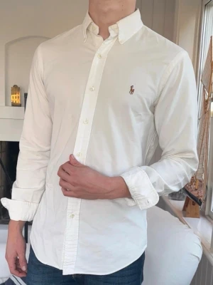 Vit skjorta från Ralph Lauren - Säljer en klassisk vit skjorta från Ralph Lauren med button-down krage och broderad logga på bröstet. Skjortan är tillverkad i 100% bomull och har långa ärmar samt knappar framtill. Perfekt för dig som gillar stilrena och tidlösa plagg.