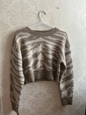 Beige och vit stickad zebramönstrad tröja - Supermysig stickad tröja i beige och vitt med zebramönster. Croppad modell med bred ribbad kant vid midjan och ärmslut. Rund halsringning och långärmad. Perfekt för dig som gillar djurmönster och vill ha en chill look.