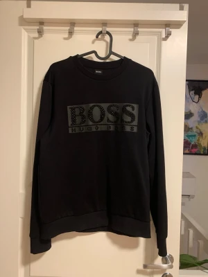Svart sweatshirt från Hugo Boss - Svart sweatshirt från Hugo Boss med stort BOSS-tryck och dekorativa detaljer på bröstet. Tröjan har rund hals, ribbade muddar och är långärmad. Materialet är mjukt och känns som bomull med inslag av polyester. Perfekt för dig som gillar stilrena och exklusiva plagg. Tröjan används en gång på en fest och räknas som en ny .