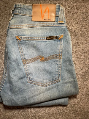 Nudie Jeans W28 L30 - Snygga ljusblå jeans från Nudie Jeans med klassisk femficksdesign och orangea sömmar. Jeansen har en rak passform och är tillverkade i mjukt bomullsmaterial. W28L30