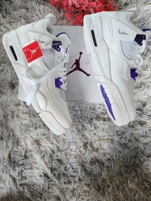 Nike Air Jordan 4 vit/lila sneakers - Nike Air Jordan 4 sneakers i vit och lila med snygga detaljer och Jumpman-logga på hälen och sulan. Skorna har snörning, platt sula och är tillverkade i skinn med meshpaneler för extra ventilation. Perfekt för dig som vill ha en ikonisk och stilren sneaker.