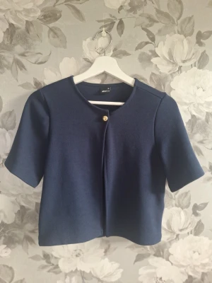 Marinblå kofta från Gina Tricot XS - Snygg marinblå kofta från Gina Tricot i storlek XS. Koftan har korta ärmar, rund halsringning och stängs med en guldfärgad knapp framtill. Materialet är mjukt och har en lätt struktur, perfekt för lager på lager under vår och höst.