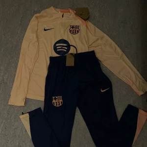 Barca träningsset - FC Barcelona träningsset från Nike med långärmad tröja och byxor. Setet är i ljusbeige och marinblått med rosa detaljer och klubbmärke på både tröja och byxa.