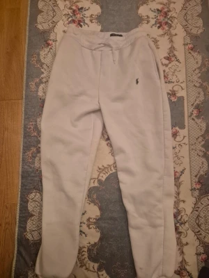 Vita mjukisbyxor från Polo Ralph Lauren - Snygga vita mjukisbyxor från Polo Ralph Lauren med klassisk broderad logga på benet. Byxorna har snörning i midjan och är gjorda i mjukt bomullsmaterial. Perfekta för chill eller häng med kompisar. Ribbade muddar vid benslut för extra komfort.
