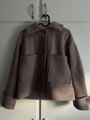 Beige ulljacka från Kappahl S - Säljer en beige jacka från Kappahl i ullblandning, storlek S. Jackan har stora fickor framtill, klassisk krage och knäppning med knappar. Ärmarna är långa med snygga manschetter och modellen är boxig och lite kortare.