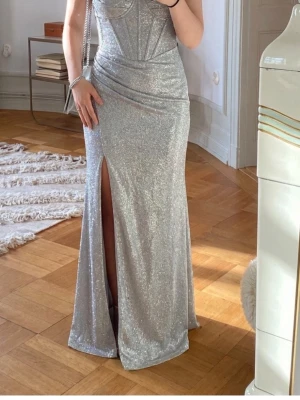 Glittrig silver långklänning med slits - Säljer en glittrig silverfärgad långklänning med snygg slits framtill. Klänningen har en figurnära passform och markerad midja, samt en snygg korsettliknande överdel. Perfekt för dig som vill sticka ut med en glamorös look.