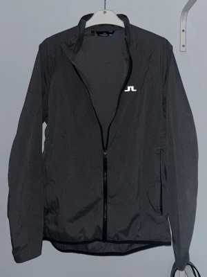 Svart vindjacka från J.Lindeberg - Svart vindjacka från J.Lindeberg med stilren design och vitt logotryck på bröstet och nacken. Jackan har hel dragkedja framtill, långärmade ärmar och praktiska fickor. Perfekt för dig som gillar sportig och clean look.