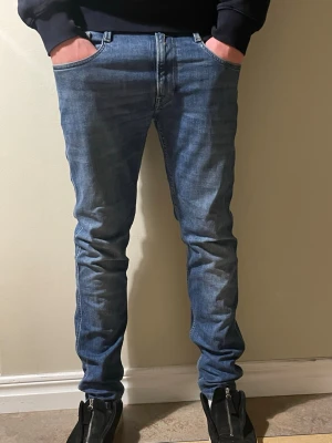Replay Anbass blå jeans herr - Säljer ett par snygga Replay Anbass jeans i klassisk blå tvätt. Modellen har smal passform, fem fickor och coola detaljer som Replay-logga på fickan. Jeansen är midwaist och har raka ben med lätt tapered snitt. Perfekta för dig som gillar stilrena och moderna jeans.