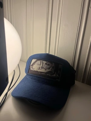 Blå mocka från Depadati - Säljer en blå mocka keps från Depadati med mesh på baksidan och justerbar rem. Framtill finns ett svartvitt tryck med motiv av när kungen blev tagen av fartkamera. Kepsen har klassisk böjd skärm och logga på insidan. Perfekt för dig som gillar streetwear.