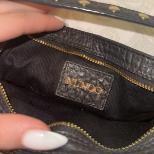 Svart axelväska från Núnoo i skinn - Säljer en svart axelväska från Núnoo i strukturerat skinn med guldfärgade detaljer och flera dragkedjeförsedda fack. Väskan har två små fickor framtill och justerbar axelrem med nitar. Perfekt för dig som vill ha en cool och praktisk väska.