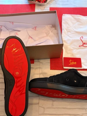 Svarta sneakers från Christian Louboutin - Säljer ett par svarta sneakers från Christian Louboutin i mocka med ikonisk röd sula. Skorna har rund tå, snörning och diskreta metallhål på sidan. Kommer med originalkartong och dustbag. Perfekta för dig som vill ha något exklusivt och stilrent.