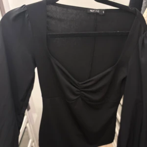 Svart blus med puffärm från NYONE - Säljer en svart blus från NYONE i storlek S. Blusen har långa puffärmar och en snygg rynkad detalj vid bysten. Modellen har en fyrkantig ringning och är figurnära i passformen. Perfekt att styla med jeans eller kjol.