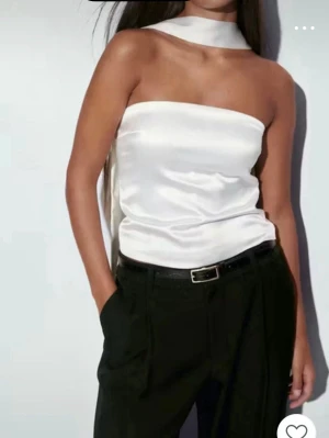 Vit bandeau tubtopp i satin - Säljer en stilren vit bandeau tubtopp i glansigt satinmaterial. Toppen är axelbandslös framtill och har en bred, lång scarf-detalj som kan knytas runt halsen eller hänga ner baktill. Perfekt för dig som gillar minimalistisk och trendig stil.