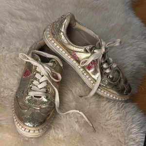 Glittriga sneakers med hjärtan och stjärnor - Säljer ett par unika sneakers i metallic guld med glittriga rosa hjärtan och silverstjärnor. Skorna har vit snörning, rund tå och platt sula. Materialet är skinn med en glittrig finish och detaljer i syntet. Perfekt för dig som gillar att sticka ut med färg och glitter.