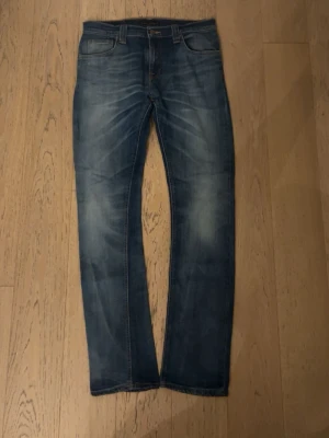 Blå jeans från Nudie Jeans Co - Säljer ett par snygga blå jeans från Nudie Jeans Co med klassisk femficksdesign och slitningar på framsidan. Jeansen har raka ben och orangea sömmar, samt Nudie-logga på bakfickorna. Perfekt för dig som gillar en avslappnad och tidlös stil. Storlek 29/32