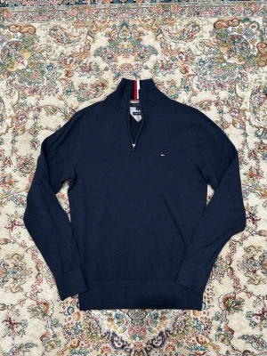 Marinblå half zip tröja Tommy Hilfiger - Snygg marinblå tröja från Tommy Hilfiger i premium bomull. Half zip med ståkrage och klassiska Hilfiger-ränder i rött och vitt på kragen. Liten logga på bröstet och ribbade muddar. Perfekt för dig som gillar stilrena och sportiga plagg.