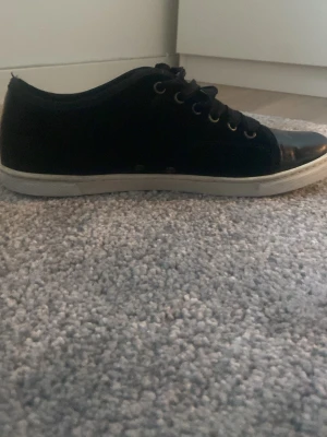 Svarta sneakers från Lanvin - Svarta sneakers från Lanvin med vit platt sula och snörning. Skorna har en rund tå och är tillverkade i skinn med en blank finish på tån. Klassisk och stilren design med metallöglor och diskret logga på sulan.