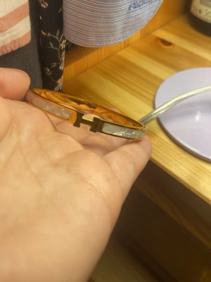 Guldfärgad armband med H-detalj - Snyggt guldfärgat armband med vit pärlemorliknande inslag och en H-formad detalj i mitten. Armbandet har en stilren och elegant design som passar perfekt till att lyfta din outfit. Perfekt accessoar för dig som gillar klassiska smycken med en modern twist.