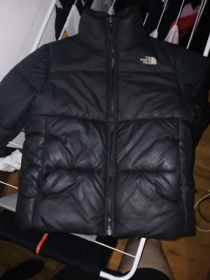Svart pufferjacka  The North Face (dam) - Säljer en svart pufferjacka från The North Face med hög krage och dragkedja framtill. Jackan har en klassisk quiltad look och är perfekt för kyliga dagar. Loggan sitter på bröstet och jackan har en riktigt clean och stilren vibe. Luva saknas därav priset