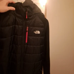 Svart pufferjacka från The North Face XL - Svart pufferjacka från The North Face i storlek XL. Jackan har huva, hel dragkedja och en bröstficka med rosa dragkedja. Perfekt för kyliga dagar och har en sportig look med quiltad framsida och logga på både fram- och baksida.