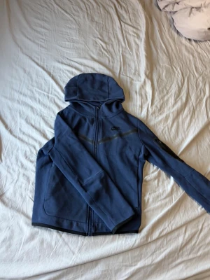 Blå Nike tech fleece  - Blå Nike tech fleece med svarta detaljer och ett snyggt Nike-logotryck på bröstet. Materialet är polyester och jackan har långärmade ärmar samt praktiska fickor. Perfekt för dig som gillar sportig stil.