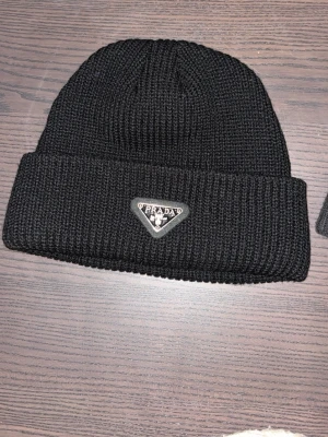 Svart  mössa från Prada helt ny  - Snygg svart mössa från Prada med klassisk ribbstickad design och uppvikt kant. Mössan har en ikonisk metallbricka med Prada-loggan framtill. Perfekt accessoar för att höja din streetstyle. Finns i flera färger  mycket bra kvalité fastpris 350kr 