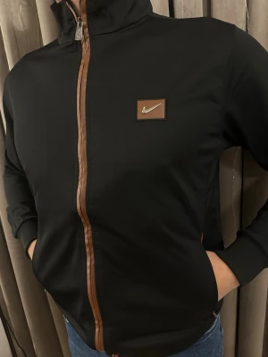 Svart Nike bomberjacka med bruna detaljer - Svart bomberjacka från Nike med bruna detaljer längs dragkedjan och ett brunt Nike-märke på bröstet. Jackan har hög krage, ribbade muddar och två sidofickor. Perfekt för dig som gillar sportig och clean stil.