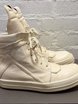 Rick Owens Geobaskets SS25 Triple Milk - Säljer ett par höga sneakers från Rick Owens i beige skinn med rund tå och platt sula. Skorna har snörning framtill, dragkedja på sidan och en chunky siluett med vadderad plös och krage. Perfekt för dig som gillar statement pieces och unik design.