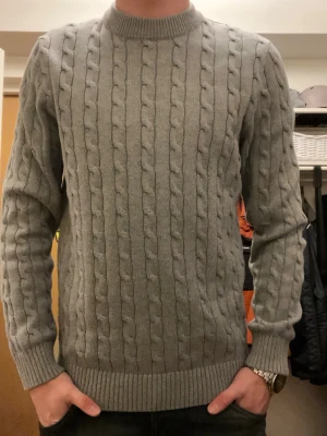 Grå kabelstickad tröja Selected Homme - Snygg grå kabelstickad tröja från Selected Homme i storlek M. Tröjan har rund hals, ribbade muddar och ett klassiskt kabelmönster som ger en stilren look. Perfekt för dig som gillar enkel men trendig stil.