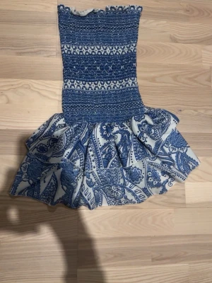 Blåvit kortklänning med volang - Supersöt kortklänning i blått och vitt med smockad överdel och volangig kjol. Klänningen har ett paisleyinspirerat mönster och är ärmlös. Perfekt för sommarens alla tillfällen! Materialet känns som bomull med inslag av syntet för stretch.